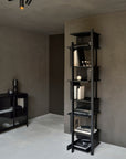 The Ethnicraft Abstract Column, a tall modern black bookshelf with asymmetrical shelves, stands against a light gray wall in a minimalist room. It holds books and decor, while a nearby black cabinet with glass doors complements the scene.
