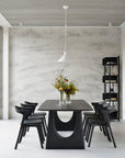 A minimalist dining room features an Ethnicraft black table with six chairs, a white pendant light above, and two flower vases. Shelving displays decor against a textured white wall beside the Ethnicraft Abstract Column.
