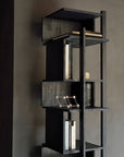 The Ethnicraft Abstract Column is a modern, asymmetrical black bookshelf crafted from FSC-certified teak, shown against a gray wall with books, a geometric object, and a small candle on its shelves.