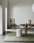 A minimalist room features a wooden chair, light gray wall, wooden table with pitcher and cups, round white stool, and the Agra Rug - Artichoke by Armadillo&Co on a concrete floor. Soft natural light streams in from the left.