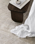 A close-up of the Armadillo&Co Agra Rug - Haze highlights its artisanal texture, complemented by a dark wooden side table with books, a decorative object, and a white fringed cloth draped over the edge.