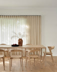 A minimalist dining room featuring an Ethnicraft Air Dining Table in oak, six matching chairs, a terra cotta vase with white dried flowers, a black apple bowl, sheer beige curtains, and light wood flooring.