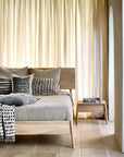 A cozy bedroom features a wooden bed, gray and black patterned cushions, a textured blanket, and Ethnicraft’s Air Nightstand—oak finish—holding books. Light curtains diffuse sunlight onto the pale wood floor for a calm setting.