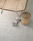 A woven chair, round wooden stool, clear glass cup, and small plate with dried flowers rest on the Andes Rug - Parchment by Armadillo&Co, its handmade wool texture elevating the light gray carpeted space.