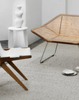A modern, minimalist room with a white sculptural vase on a small round table, a woven rattan lounge chair with metal legs, a wooden chair with a white seat, and an Armadillo&Co Andes Rug - Parchment for textured charm.