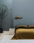 A modern minimalist bedroom with muted blue walls, an olive green bedspread, matching pillows, a handmade Andes Rug - Parchment by Armadillo&Co, a potted tree, a stone side table, and decorative objects on a sleek headboard shelf.