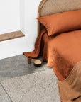 A bed with burnt orange linen bedding and a wicker headboard sits on a stone floor next to the Andes Rug - Rye by Armadillo&Co. Nearby are a small brown cup and wooden bowl. A wooden shelf is mounted on the white wall.