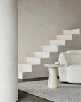 Minimalist interior with a floating white staircase, round marble accent table, textured white sofa, large column, and neutral walls and carpet layered with Armadillo&Co’s Andorra Rug - Albatross, renowned for its 100% wool handmade artisan quality.