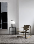 A modern, minimalistic room featuring a bronze rocking chair, a small black side table with a white lamp, an Armadillo&Co Andorra Rug - Pepper, and a large abstract black-and-gray painting against a light gray wall.