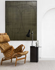 A modern interior features a tan leather lounge chair, a black side table with a lamp and geometric decor, and an Andorra Rug - Terrazzo by Armadillo&Co beneath a large abstract painting with black lines on muted green on a white wall.
