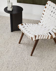 A modern chair with wooden legs and a white woven seat stands by a small black side table topped with a white abstract sculpture, all placed on the Andorra Rug - Terrazzo by Armadillo&Co, crafted from Fair Trade wool.