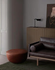 A modern living room features a dark Natadora leather sofa, a round Fable Ottoman by Wooden Stories, a ribbed sideboard, a table lamp, and a portrait of a woman in historical attire. Sunlight streams through tall white French doors.