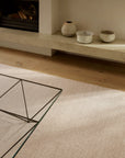 An Armadillo&Co Aspen Rug - Sandstorm, a hand-knotted, beige wool rug, in a room with a fireplace, stone hearth, and glass-topped coffee table.