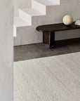 A minimalist interior with textured white stairs, a dark wooden bench with a round beige object, and the Aspen Rug - Natural by Armadillo&Co—hand-knotted from 100% wool—on the floor, set against a rough off-white wall.