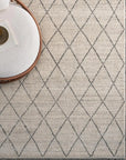An Atlas Rug in Limestone by Armadillo&Co with a diamond lattice design lies on a speckled concrete floor. The upper left shows part of a round, light chair and a small table.