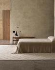 A minimalist bedroom features beige textured walls, a bed with a beige blanket, a wooden side table, and a bench displaying a modern black sculpture. The Atlas Rug - Limestone by Armadillo&Co anchors the space next to the closed wooden door.