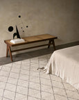 A minimalist bedroom with a wooden bench, books, a black sculpture, and a bed in beige linen. The floor showcases the Armadillo&Co Atlas Rug in Limestone with a subtle grid pattern, set against textured neutral walls.