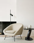 The Ethnicraft Barrow Lounge Chair - Off White sits beside a round black side table with a white teapot and mug, complemented by a minimalist black floor lamp, set against a neutral wall and light rug for a modern touch.