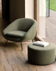 The Barrow Lounge Chair - Pine by Ethnicraft and its matching ottoman sit on a wood floor near a large window, topped with books. Green grass and brick pavement are visible outside, enhancing the space’s subtle vintage vibe.