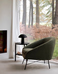 A Barrow Lounge Chair - Pine by Ethnicraft sits beside a small round table with a black lamp, near a large window overlooking forest views filtered through sheer curtains and tall ferns.