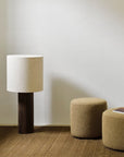 A modern floor lamp with a dark wooden base and white shade stands next to two Ethnicraft Barrow Pouf - Ginger / Small, upholstered in Italian fabric, on a brown textured rug against a plain white wall.