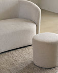 A modern, light beige curved sofa and the Ethnicraft Barrow Pouf - Off White / Small rest on a textured beige rug in a minimalist room with neutral-toned walls and flooring.