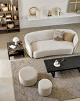 A modern living room with a curved beige sofa, two Ethnicraft Barrow Poufs in Off White (Large), a black coffee table, a side table with lamp, neutral-toned decor on shelves and walls, all atop a textured beige rug.