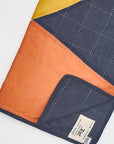 The FAIRE/ANCHAL Sunset Quilt features geometric navy, orange, and yellow color blocks with white grid stitching. Hand-stitched from organic cotton, it has a sewn label on the lower corner and is set against a plain background.