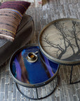 Two round side tables from Ethnicraft’s Tray Collection feature metal frames; one is topped with the large Black Tree Wooden Tray, while the other tray shines blue with espresso. Nearby, a gray chair with a patterned pillow evokes European refinement.