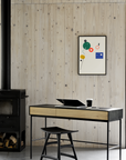 A minimalist workspace features Ethnicrafts Blackbird Desk with a laptop and glass, a black chair, abstract art on a light oak-paneled wall, and a black wood stove with logs neatly stacked below.