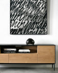 An Ethnicraft Blackbird TV Cupboard with books and a dark abstract sculpture is placed beneath a large black-and-white artwork of bold, overlapping calligraphic script on a white wall.