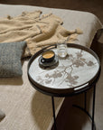 The Ethnicraft Blossom Wooden Tray - Round / Small, featuring a floral design, holds a black cup of coffee and a glass of water beside a neatly made bed with a textured beige blanket and blue-gray cushion: classic European refinement.