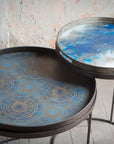 Two round metal side tables from Ethnicraft, each topped with the Blue Mist Organic Glass Tray (Round/Small): one tray shows a blue and gold circular pattern, the other a blue and white abstract design, both on slim black tripod legs.