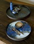 An Ethnicraft Blue Mist Organic Glass Tray (Round, Small) sits on a wooden table, displaying blue ceramic items; another tray features a blue and white marbled pattern with stacked plates and cutlery, evoking European refinement.