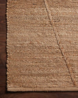 A close-up of the Bodhi Rug - Natural / Natural Abstract by Loloi, a 100% jute rug with textured natural fibers on a dark wood floor. A thick diagonal strand contrasts with the horizontal weave.