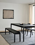 A minimalist dining room features a black varnished table, the Ethnicraft Bok Bench in Black Oak, and a black chair on a gray rug. Beige curtains cover the window while a blank textured frame hangs on the white wall, filled with natural light.