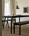 Minimalist dining room with a black wooden table, matching chairs, and the Ethnicraft Bok Bench in Brown Oak set on a woven rug. Soft beige curtains frame tall windows, complemented by neutral decor and abstract art for a calm atmosphere.