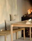 The Ethnicraft Bok Bench - Oak with beige cushions sits beside a matching dining table, topped with a teapot and pastries. A cozy fireplace and stacked firewood complete the warm, minimalist setting.