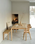 Minimalist dining area with an Ethnicraft solid oak table, matching chairs, and the Bok Bench - Oak. A vase with a branch decorates the table. Cozy fireplace in corner, snowy views visible through a large window.