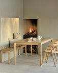 A neutral, softly lit dining area features an Ethnicraft Bok Bench - Oak, paired with a solid oak table and chairs from the same collection, light-toned cushions, a corner fireplace aglow, and stacked firewood below.