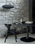 A modern workspace with a round black table, an Ethnicraft Bok Dining Chair in black oak, a laptop, books, and bottled water set against a stone wall under a black disc pendant light.