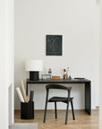 Minimalist home office with a black desk, Ethnicraft Bok Dining Chair in Black Oak, and matching lamp. Art supplies and decor are neatly arranged, with rolled papers in a black container. Abstract art hangs on the white wall.