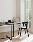 A minimalist dining area with a solid oak table, Ethnicraft Bok Dining Chair in Black Oak, neutral bench cushions, and a glass pitcher with dishes. Sunlight streams through a large window overlooking the garden.