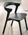 A Bok Dining Chair - Black Oak by Ethnicraft, featuring a curved backrest and minimalist design, sits on a light wood floor beside a light-colored table.