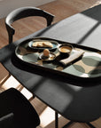 A modern black dining table with a patterned tray holds breakfast items, as sunlight creates soft shadows on the wooden floor and an Ethnicraft Bok Dining Chair in Black Oak.