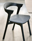 The Ethnicraft Bok Dining Chair in Black Oak with Grey Fabric features a curved open backrest and cushioned seat, shown here on a light wood floor beside a light-colored table.
