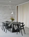 Minimalist dining room with Ethnicraft Bok Dining Chairs in black oak with leather around a black table, modern white pendant light, textured gray wall, cream floor-to-ceiling curtains, and a vase of colorful flowers as centerpiece.