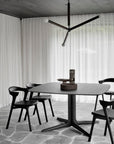 A minimalist dining area features a dark solid oak table, four Ethnicraft Bok Dining Chairs in Black Oak with Leather, a sculptural centerpiece, stone tile flooring, sheer white curtains, and a modern black chandelier above.