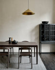 A minimalist dining room featuring a dark wood table, four Ethnicraft Bok Dining Chairs in Brown Oak, a gray rug, a cabinet with black dishware, and a gold pendant light against a plain light wall.