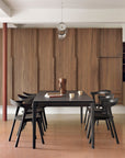 A modern dining room with a black rectangular table, six Ethnicraft Bok Dining Chairs in brown oak, a decorative wooden vase, three cups on the table, wood-paneled walls, and a minimalist pendant light overhead.